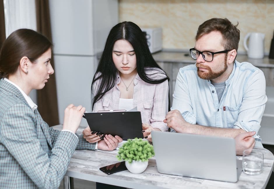 A mortgage loan officer reviewing documents with a couple over a laptop at a kitchen table.